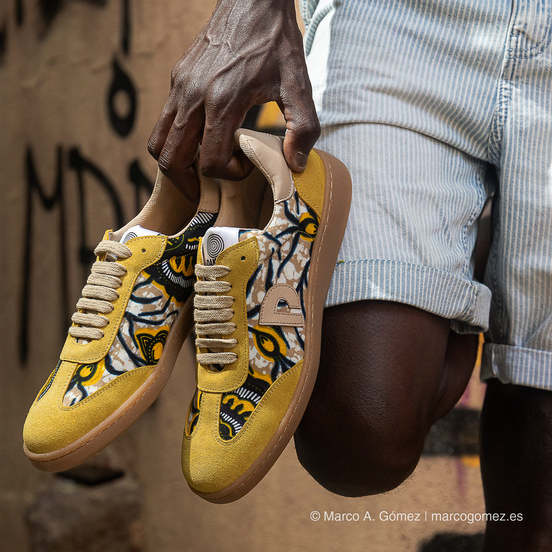 Proyecto integral Manos sosteniendo zapatillas de lona amarilla con patrones tradicionales africanos de la marca Dakart por Marco A. Gómez.